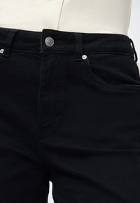 Čierne riflové nohavice s hladkou textúrou, zapínaním na gombík a striebornými kovaniami. Majú predné vrecká a púzdra na opasok.