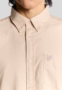 Beige overhemd met knopen en een kraag, voorzien van roze knopen en een klein geborduurd logo op de borstzak. Geweven stof.