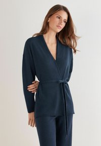 Marineblaue Wickelstrickjacke aus weichem Strickmaterial, mit V-Ausschnitt, langen Ärmeln und gebundenem Bund. Schlichte Textur und klare Linien.