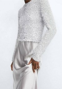 Pullover cropped argento con una texture, maniche lunghe abbinato a una gonna in satin grigio chiaro, mettendo in evidenza il contrasto dei tessuti e la lucentezza.