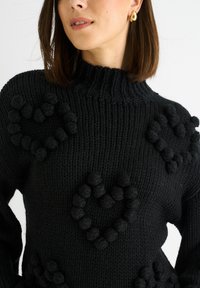 Maglione nero a maglia con collo alto, caratterizzato da cuori pom-pom testurizzati disposti in gruppi sul davanti. Materiale spesso e caldo con finitura a coste.
