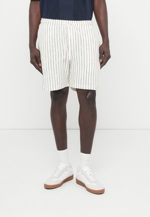Shorts rayés blancs avec une taille élastique, coupe décontractée, matière texturée, portés avec des baskets blanches sur un fond uni.