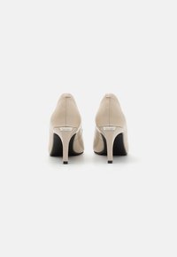 Beige bőrsarkú cipők hegyes orral, fekete talppal és vékony stiletto sarkakkal. Diszkrét márkázás a hátsó részen. Simább tapintás.