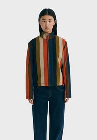 Giovane donna in piedi, che indossa una giacca con zip a righe verticali multicolori e jeans blu scuro, su uno sfondo chiaro e semplice.