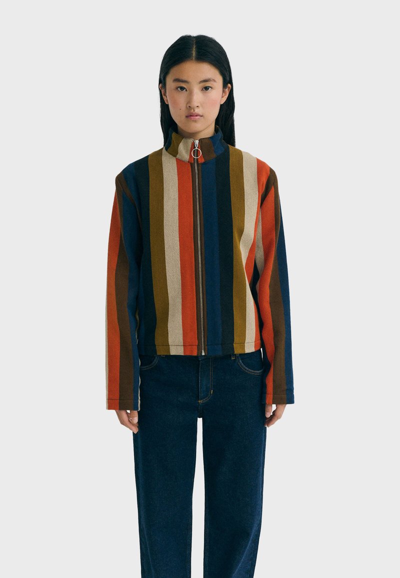 Giovane donna in piedi, che indossa una giacca con zip a righe verticali multicolori e jeans blu scuro, su uno sfondo chiaro e semplice.