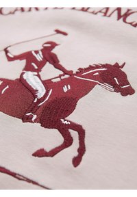 Broderie d'un joueur de polo en bordeaux et blanc, montant à cheval et balançant un maillet sur un tissu beige clair.