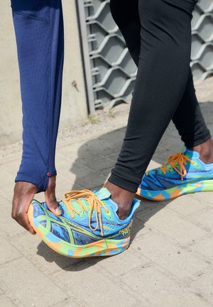 Heldere blauwe sportschoenen met kleurrijke abstracte patronen, voorzien van oranje veters en een textuurrijke zool, gecombineerd met zwarte legging.