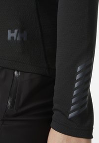 Czarny sportowy top z teksturowanej tkaniny, z subtelnym logo i paskiem na rękawie. Zawiera szczegół z kieszenią na zamek.