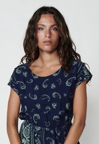 Rochie bleumarin cu decolteu rotund, mâneci scurte și talie strânsă. Modele complexe de paisley verde și flori pe întreaga suprafață.