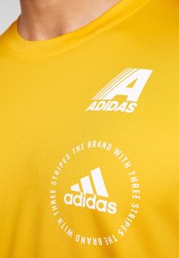 Gelbes T-Shirt mit kurzen Ärmeln aus glattem Stoff. Verfügt über weiße Adidas-Logos: ein stilisiertes "A" und eine kreisförmige Markeninschrift.