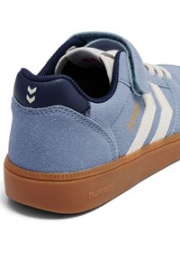 Lichtblauwe suède sneaker met een marineblauwe hiel, witte strepen en een klittenbandsluiting. Voorzien van een gestructureerde rubberen zool en geperst merklogo.