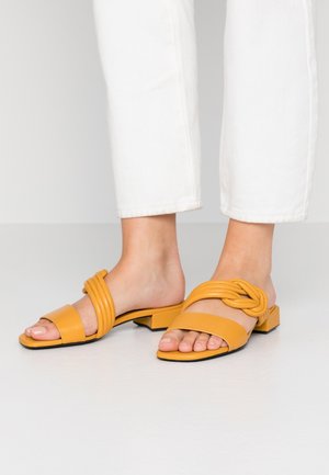 Gule slidesandaler lavet af glat syntetisk materiale med en knude på stroppen og en lav blokhæl, stylet med hvide bukser, der går til anklerne.