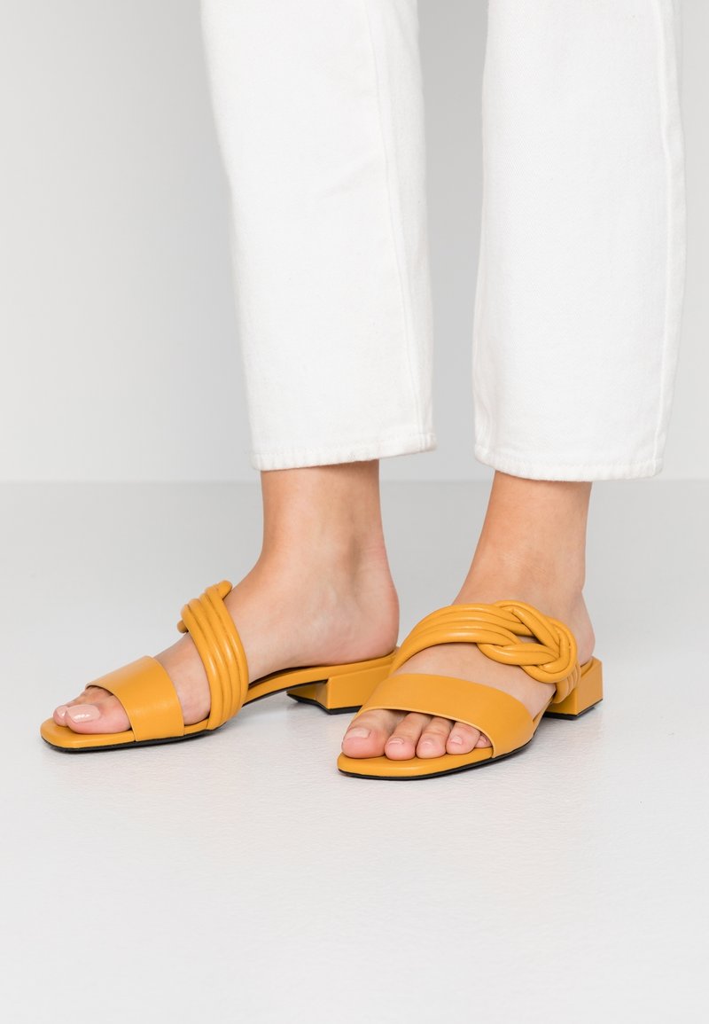 Sandales jaunes à enfiler en matériau synthétique lisse, avec une bride nouée et un petit talon bloc, associées à un pantalon blanc coupé à la cheville.
