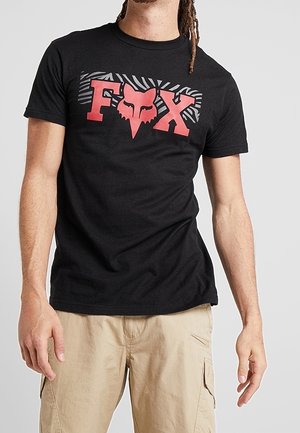 Mann trägt ein schwarzes T-Shirt mit rotem "FOX"-Schriftzug und Fuchs-Kopflogo, kombiniert mit beigen Cargo-Shorts vor einfachem Hintergrund.