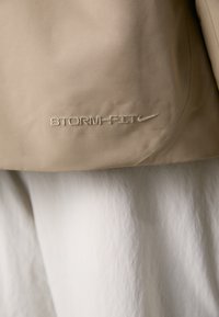 Beige waterproof jacket featuring "STORM-FIT" logo in pale thread, smooth texture, and a streamlined design. Accented with a Nike swoosh.