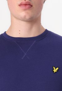 Lila kapucnis pulóver bordás nyakkal, bal mellen egy kis sárga hímzett saslogóval.