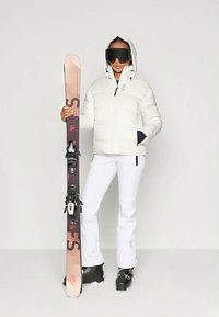 Hvid pufferjakke med hætte, sorte skibukser og sorte skistøvler. Holder en pink ski med sorte bindinger. Store sorte goggles bæres.