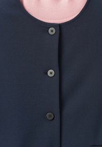 Gros plan sur un vêtement bleu marine à boutons sur un tissu ribbed rose clair, montrant trois boutons foncés sur le devant.