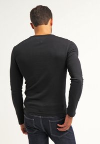 Man med kort brunt hår som bär en åtsittande svart långärmad tröja och mörkblå jeans, stående vänd bort med höger hand nära bakfickan.