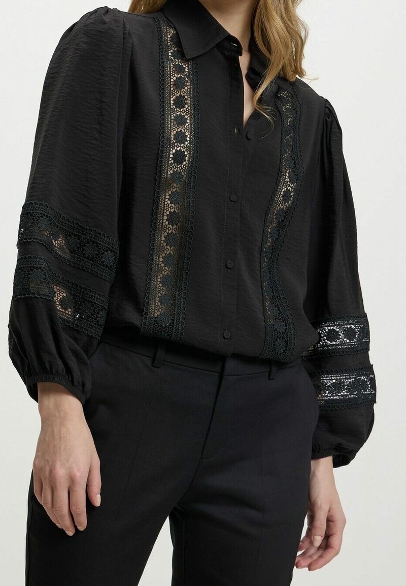 Blouse noire à manches longues boutonnée avec panneaux en dentelle florale à l'avant et aux manches, associée à un pantalon noir sur une personne debout.
