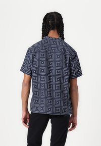 Rövid ujjú ing sötétkék anyagban, fehér pöttyös geometrikus négyzetmintával. Szabvány gallér és laza fazon.