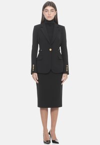 Blazer negro a medida con botones dorados, combinado con una falda negra ajustada. El conjunto incluye una blusa negra de cuello alto debajo. Zapatos de tacón negros completan el look.