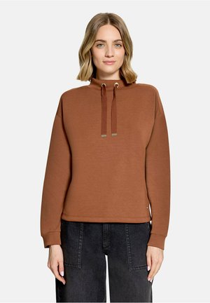 Kvinde med skulderlangt blondt hår iført en brun langærmet sweatshirt med snore og sorte højttaljede jeans mod en hvid baggrund.