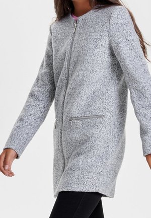 Femme portant un manteau zippé texturé gris clair mi-cuisse avec deux poches zippées, associé à un pantalon noir et une chemise rose en dessous.