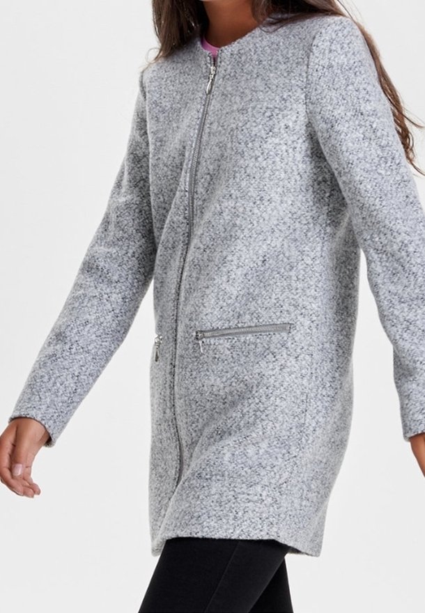 Femme portant un manteau zippé texturé gris clair mi-cuisse avec deux poches zippées, associé à un pantalon noir et une chemise rose en dessous.
