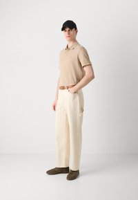 Polo beige, pantalon large crème avec poches latérales, ceinture en cuir marron et casquette noire. Des chaussures marron complètent la tenue.