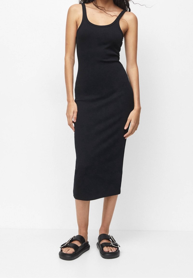 Femme portant une robe midi noire ajustée sans manches et des sandales à plateforme noires avec de grandes boucles, debout devant un fond blanc.