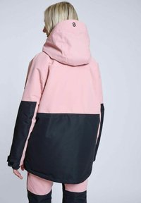 Lyserød og sort vinterjakke med hætte, der har et farveblokdesign og tekstureret stof. Bemærkelsesværdig logo på ryggen. Løs pasform.