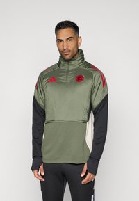 Αθλητικό μπουφάν quarter-zip σε ελιά με μαύρα μανίκια, κόκκινες λεπτομέρειες και λογότυπο του συλλόγου. Διαθέτει κομψή, λεία υφή και τρύπες για τους αντίχειρες.