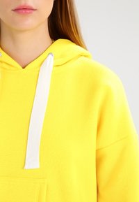 Felpa gialla con tasca frontale, dotata di un laccio bianco e una texture morbida. Il design presenta una vestibilità rilassata e un colletto con cappuccio.