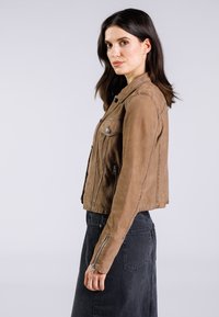 Veste en daim marron au design court, avec des quincailleries en métal argenté et des poches zippées. Comprend des coutures le long des épaules et des poignets pour plus de structure.