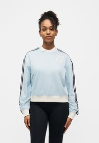 Νεαρή γυναίκα που φοράει γαλάζιο φούτερ με μαύρες ρίγες στα μανίκια και τον αριθμό 22, στέκεται μπροστά σε λευκό φόντο.