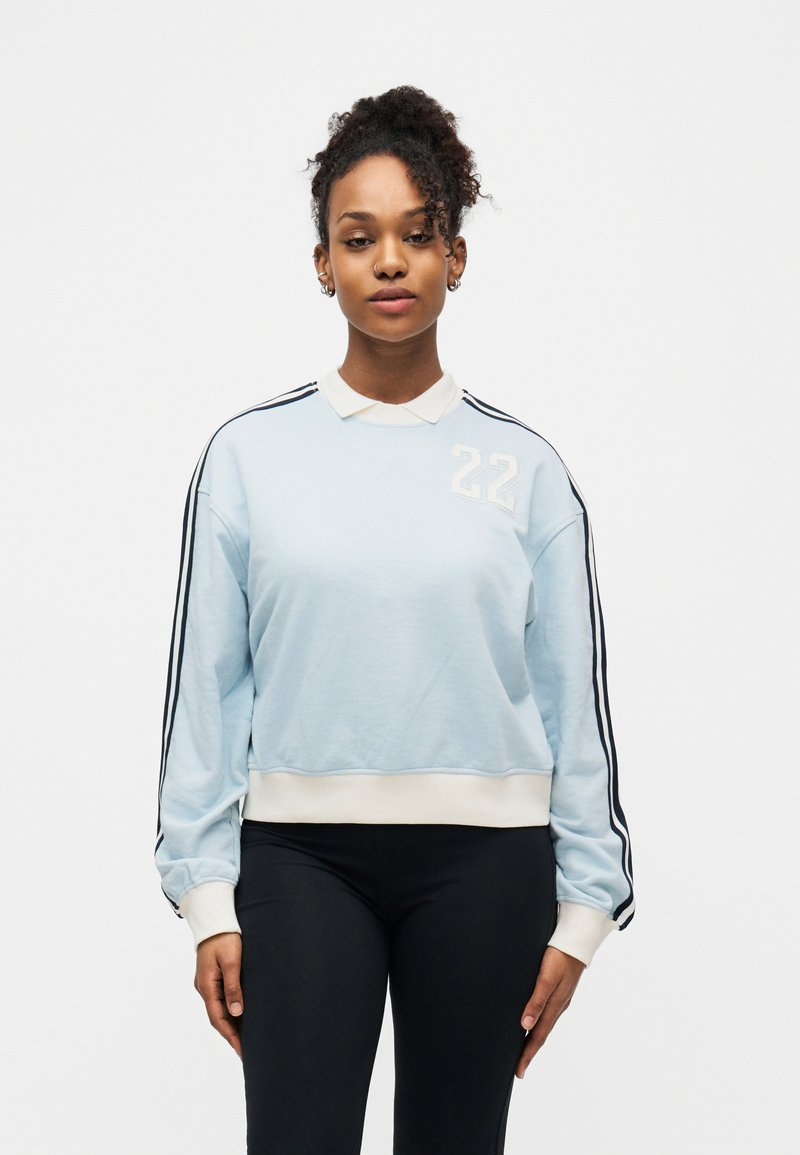 Νεαρή γυναίκα που φοράει γαλάζιο φούτερ με μαύρες ρίγες στα μανίκια και τον αριθμό 22, στέκεται μπροστά σε λευκό φόντο.