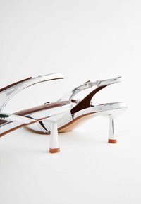 Escarpins slingback en cuir verni argenté avec talons fins inclinés et semelles intérieures marron sur fond blanc.