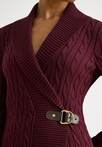 Vinrød strikket cardigan med kabelmønster, bred krage og et brunt lærbelte med gullfarget spenne som lukking.