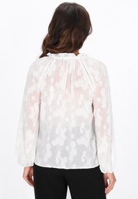 Blusa blanca con un patrón floral texturizado, mangas largas abullonadas y cuello con volantes. Material translúcido con un ajuste relajado.