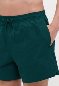 Shorts de bain vert foncé avec une taille élastique et un cordon de serrage. Tissu texturé avec des poches latérales et une petite fente à l'ourlet.