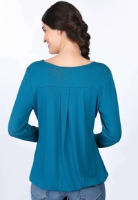 Türkises Langarmoberteil mit rundem Ausschnitt, gerafftem Rückenteil und weichem Stoff, kombiniert mit hellblauen Jeans.