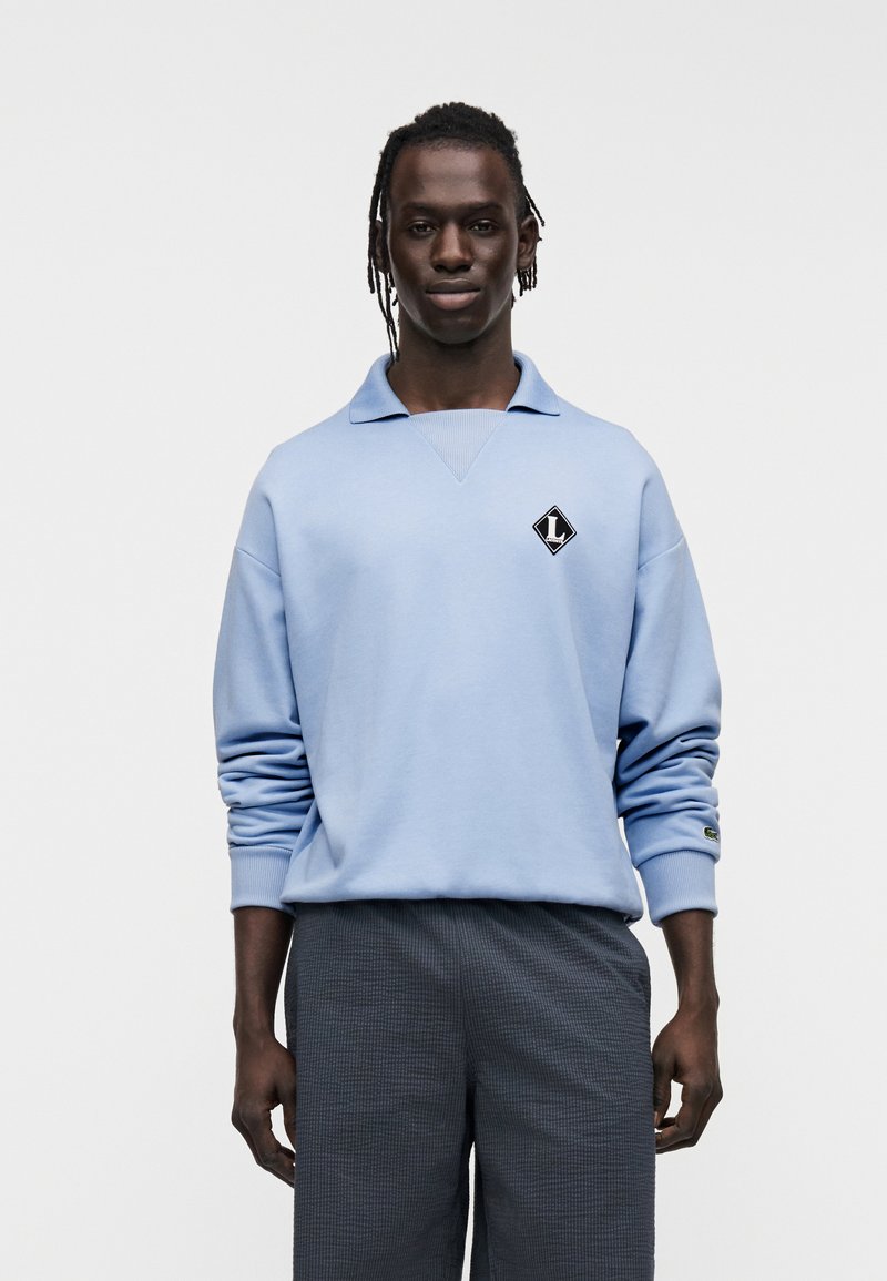 Sweat-shirt bleu clair avec col polo, arborant un logo losange noir. Porté avec un pantalon texturé gris foncé. Tissu lisse et coupe décontractée.