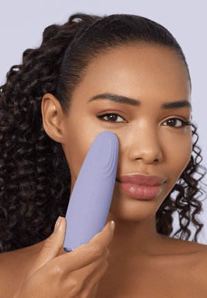 Femme aux cheveux bouclés tenant un appareil de nettoyage facial à la lavande contre sa joue droite, sur un fond violet clair.