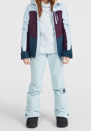 Enfant portant un pantalon de neige bleu clair, des bottes noires, un pull blanc texturé et une veste d'hiver color-block avec des panneaux bleus et violets.