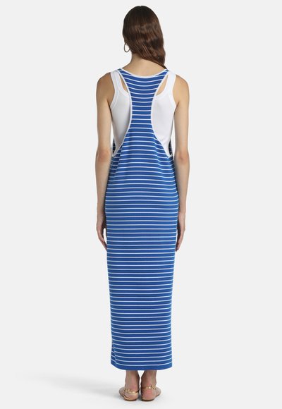 Femme debout de dos portant une robe maxi à rayures bleues et blanches avec un dos nageur et un débardeur blanc en dessous.