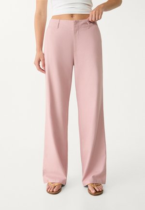 Vrouw draagt roze broek met een hoge taille en wijde pijpen en een wit cropped topje, houdt de tailleband met één hand vast, draagt beige sandalen.
