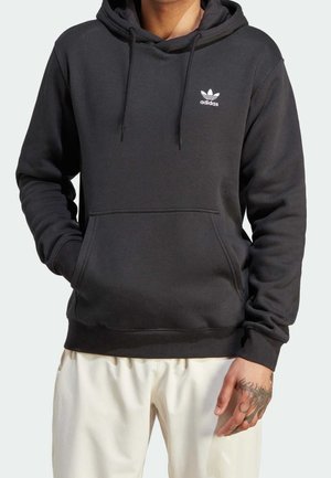 Mand iført sort Adidas hættetrøje med frontlomme og hvidt Adidas-logo på brystet, kombineret med lyse bukser og tatovering på højre hånd.