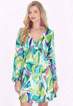 Vrouw met lang blond haar draagt een kleurrijke lange mouwen jurk met groene, blauwe, paarse en witte patronen en grote paarse tassel oorbellen.