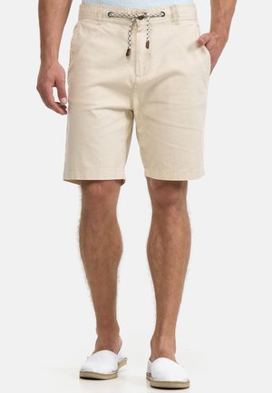 Beige Baumwollshorts mit Kordelzug in der Taille, Seitentaschen und einem lockeren Sitz. Kombiniert mit weißen Slip-On-Schuhen. Glatte Textur.